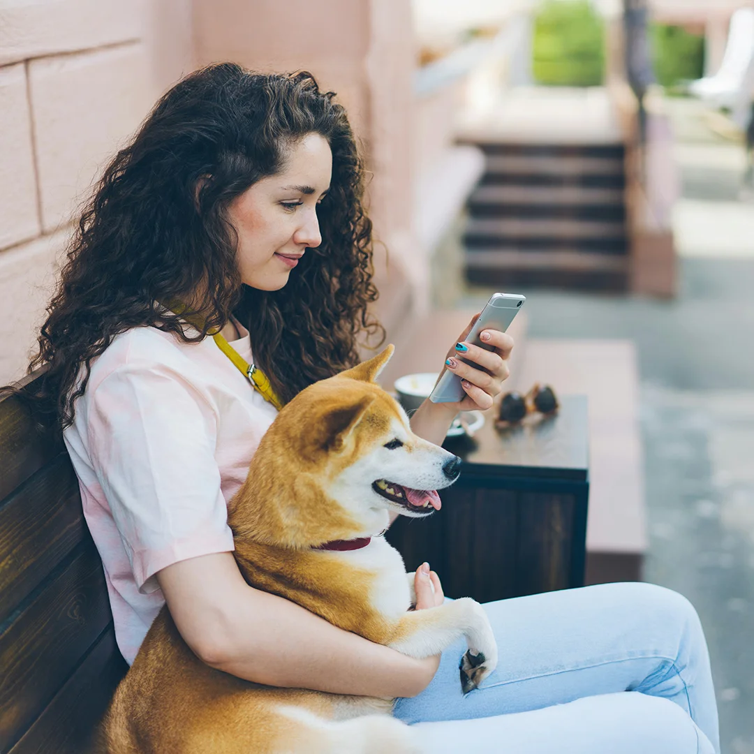 A woman sits on a bench outside, holding a Shiba Inu in her lap while looking at her smartphone.